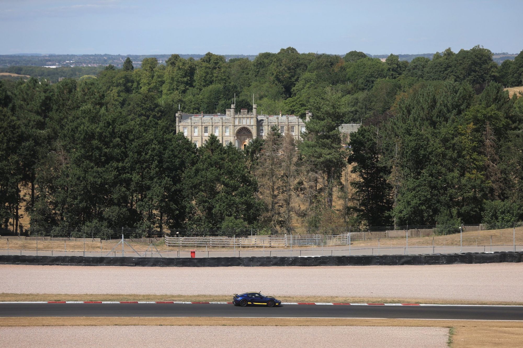 A blue Porsche GT4 on track at Donington Park, with Donington Hall in the trees behind