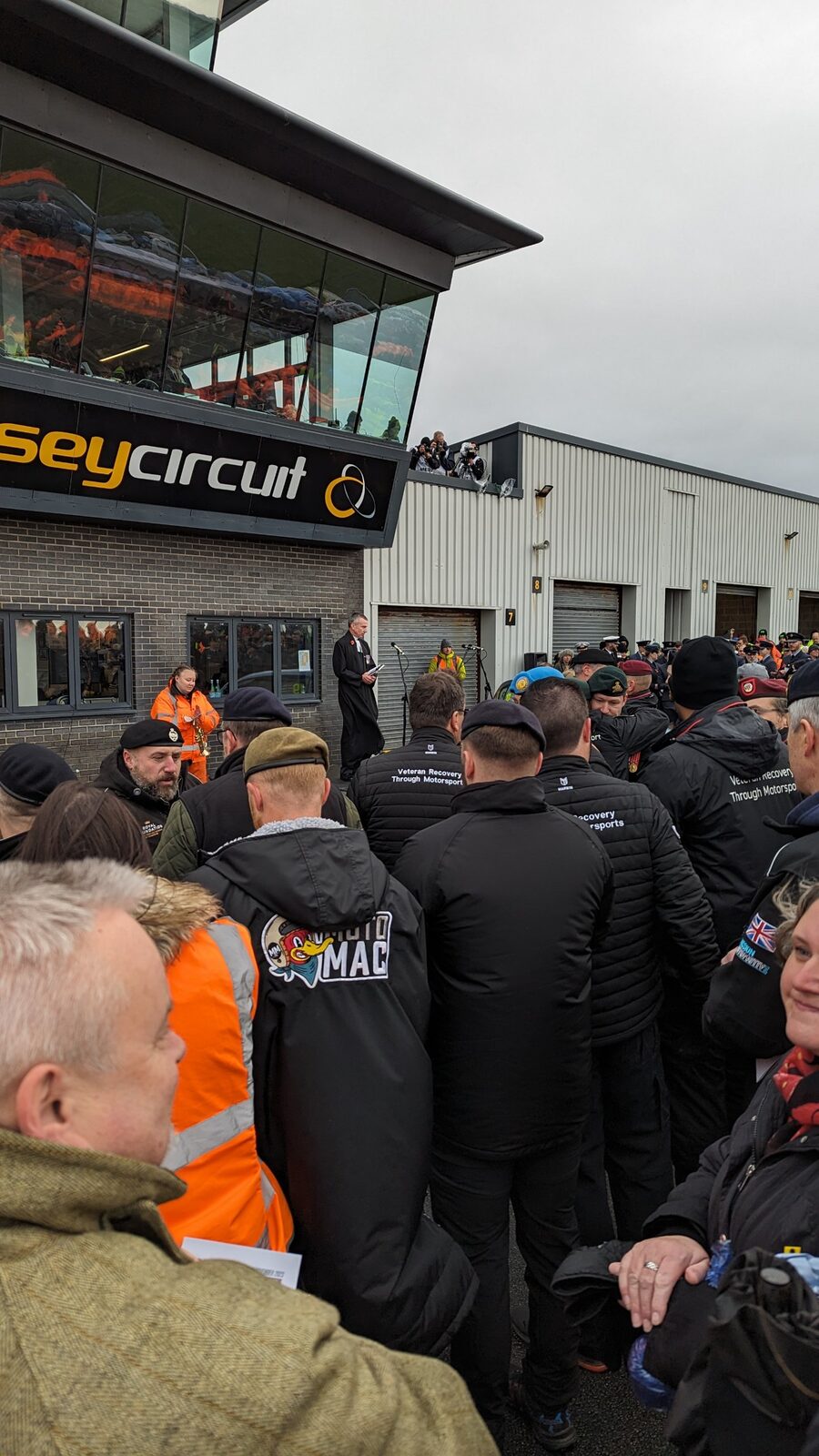 A chaplain leads the Service of Remembrance outside the control tower at Anglesey Circuit, veterans gathered in Mission Motorsport jackets.